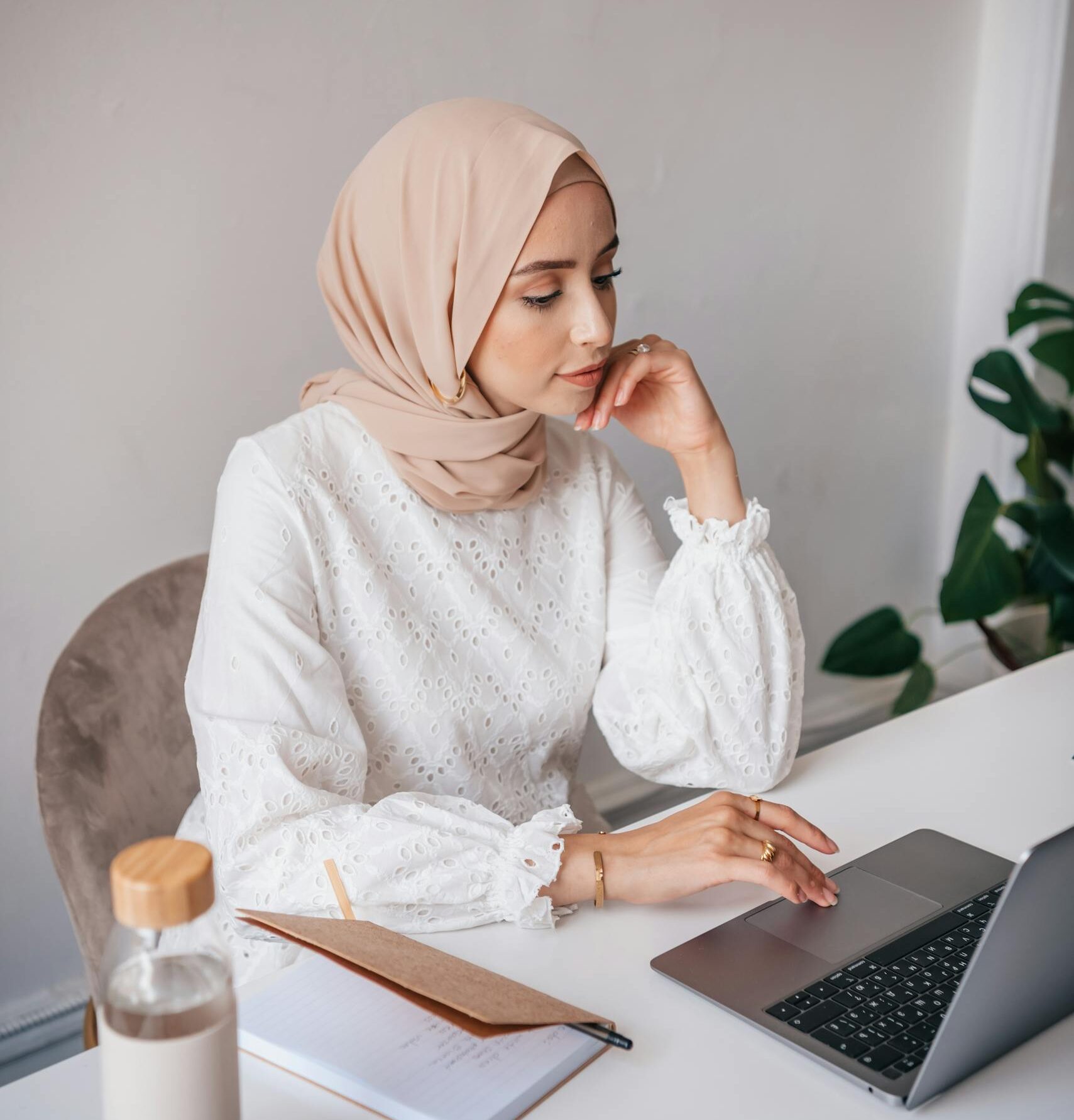Woman wearing a hijab working on a laptop at a desk in a minimalist room with a bottle and notebook.